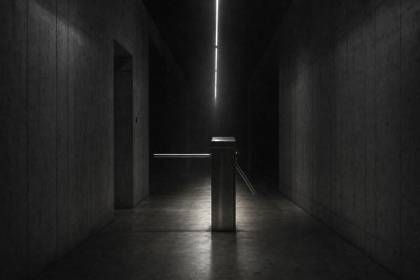 A closed steel turnstile standing alone in a dim concrete corridor, lit by a single overhead strip of light